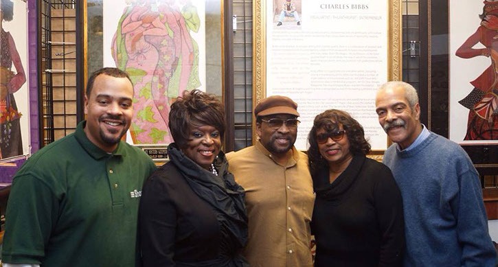 Five smiling individuals pose together at an event showcasing the artwork of Charles Bibbs, with colorful pieces displayed in the background.