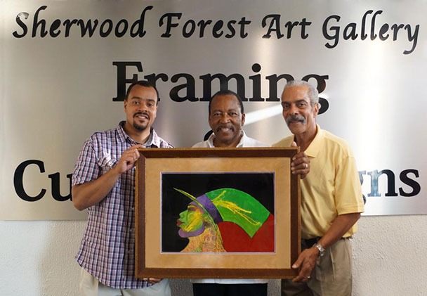 Three men pose with a vibrant framed artwork depicting a profile of a figure. They stand in front of the Sherwood Forest Art Gallery sign, showcasing community engagement in art.