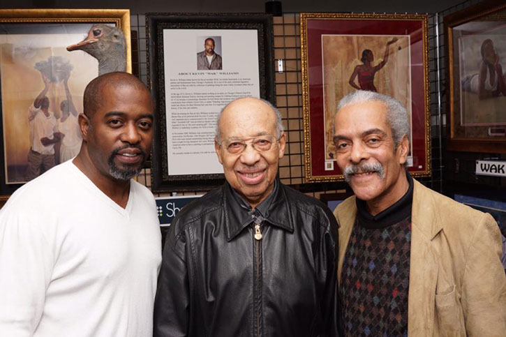 Three men pose together in front of framed artworks, smiling warmly. The central figure, an elderly man, wears glasses and a leather jacket.