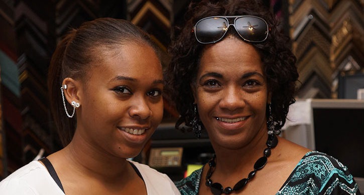 A woman and a young girl smile together in a framing store, surrounded by colorful frame samples. The image reflects their positive relationship and business context.