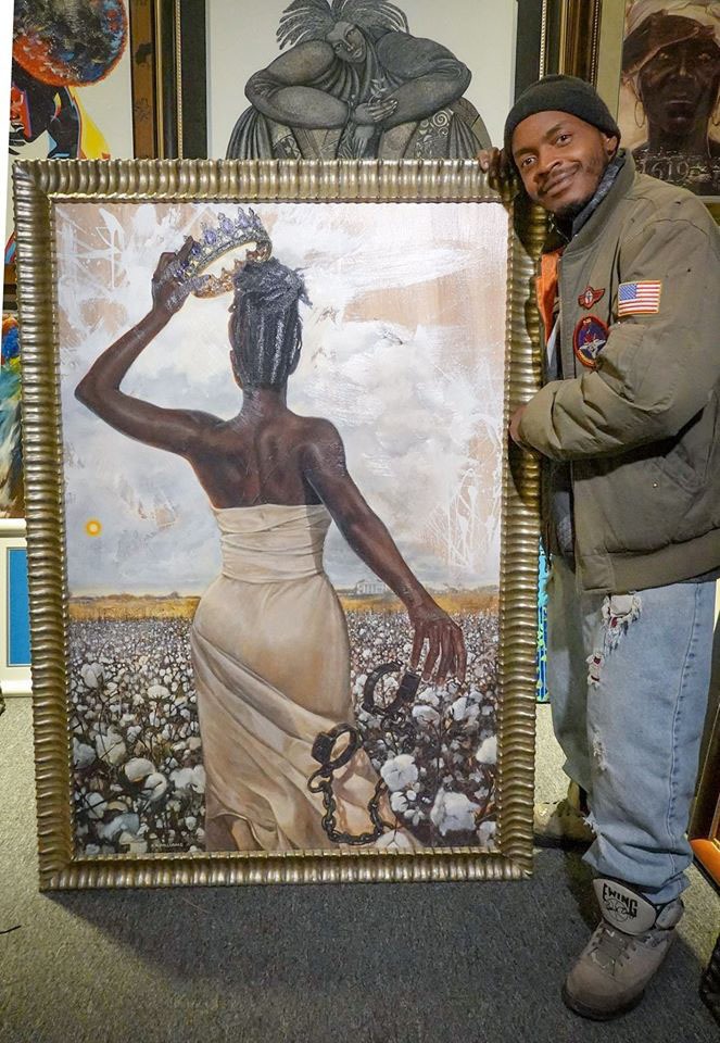 A man stands next to a framed painting of a woman in a white dress holding a crown, symbolizing liberation against a backdrop of cotton fields.