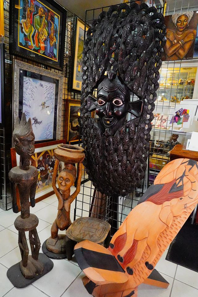 A diverse array of African sculptures and art pieces, including a large carved mask, wooden figures, and a decorative chair, showcased in an art gallery.