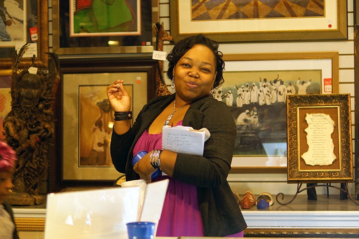 A woman with curly hair, wearing a black cardigan and purple dress, stands confidently in an art gallery, smiling and holding a notebook.