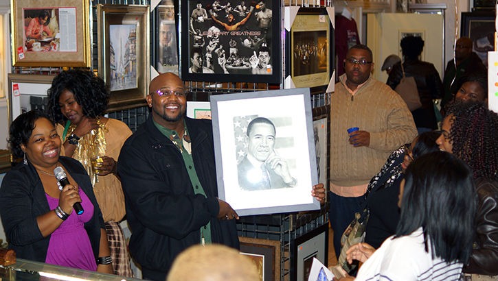 A man proudly holds a portrait of Barack Obama, surrounded by an engaged crowd in a gallery setting, highlighting a celebration of art and culture.