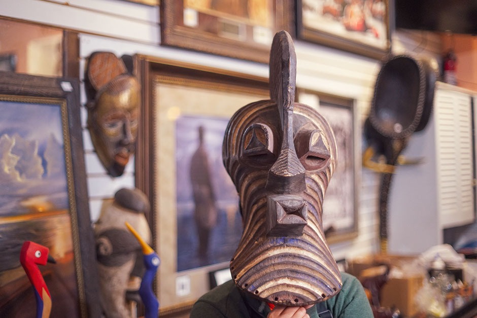 Carved wooden mask held by a person, surrounded by framed artworks and decorative items, showcasing cultural artistry and craftsmanship.