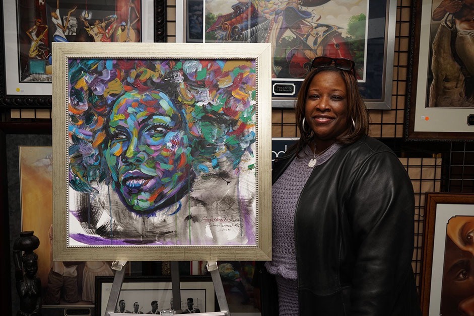 A woman stands beside a vibrant, colorful portrait of a woman with expressive features, showcasing artwork in a gallery setting.